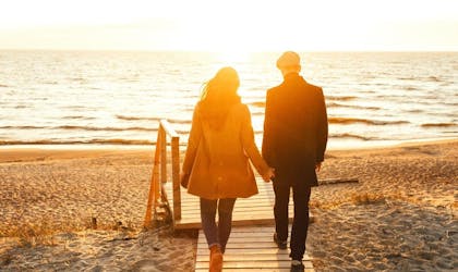 Par som holder hender går tur på strand i solnedgang