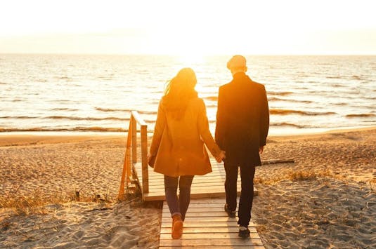 Par som holder hender går tur på strand i solnedgang